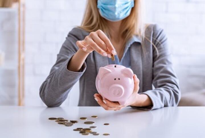 Business woman with mask puts money into piggy bank Business woman with mask puts money into piggy bank